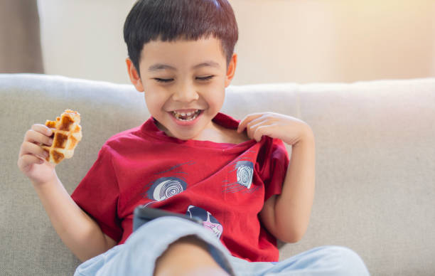 istock Cute Asian child boy laughing while eating a waffle and using smartphone at home. 2161135651
