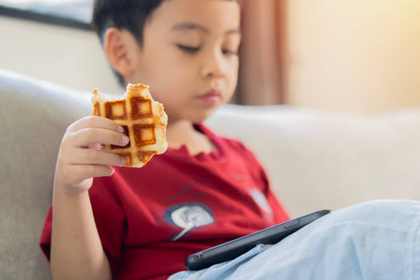 istock Little boy kid holding a waffle and playing mobile or watching video while resting at home. 2161135719