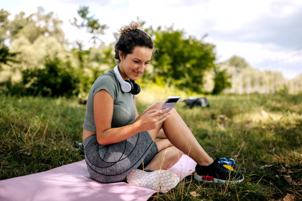 istock Young smiling woman using phone while sitting on yoga mat at public park 2161136705