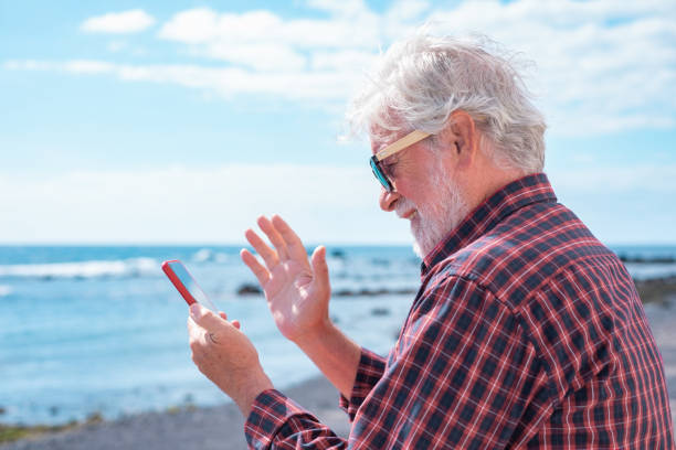 istock Smiling senior bearded man waves in mobile phone while making video call, senior pensioner enjoys nice sunny day sitting outdoors facing the sea 2161139544