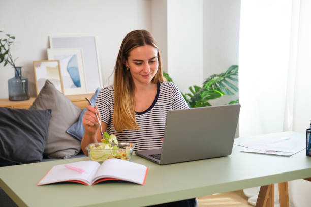 istock Young woman eating salad while working from home using laptop 2161143438