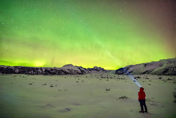 A person stands under the vibrant northern lights in Skaftafell, Iceland, holding a flashlight that illuminates the snowy ground. The green hues of the aurora borealis fill the night sky above the mountainous landscape, creating a breathtaking natural display.