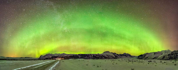 A stunning panoramic shot of the northern lights arching across the starry night sky over Iceland's snow-covered mountains. The vibrant green hues of the aurora create a striking contrast against the dark, rugged terrain and the expansive snowy landscape near Skaftafell glacier