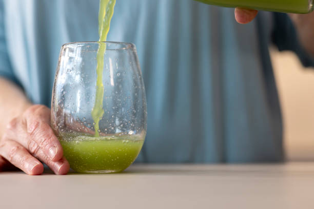 Close up of a woman standing in her kitchen preparing a green health drink. She is on a health journey. She is pouring the juice into a glass.\n\nVideos are available similar to this scenario.