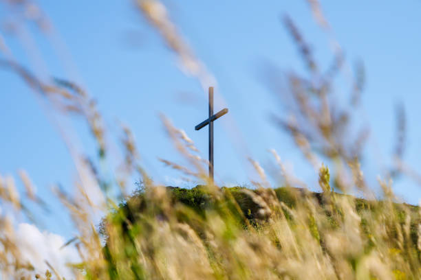 Large white cross monument positioned on rocky outcrop overlooking diverse agricultural landscape. Aerial view shows patchwork of farm fields, orchards, and rural roads near Marsing, Idaho. Rocky formation creates natural pedestal for religious marker. Distant mountains frame horizon while scattered rural homes and farm buildings dot countryside. Summer conditions highlight contrast between irrigated green fields and dry terrain.