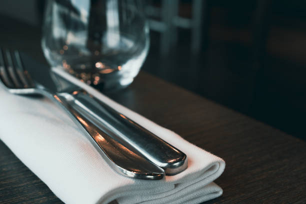 A close-up shot of a knife and fork resting on a white napkin on a dark wooden table in a restaurant. The silverware is polished and reflects light. A glass is out of focus in the background.