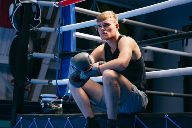 A young man wearing a sports vest and shorts sits on the edge of a boxing ring. He is wearing boxing gloves and has a determined expression on his face.