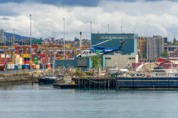 An elevated view of Vancouver reveals a mix of forested residential areas, industrial port facilities, and the downtown skyline. Cargo ships and a cruise liner are docked along the waterfront, with cranes and silos visible in the middle ground. The white sails of Canada Place stand out among high-rise buildings, while distant mountains and water bodies stretch across the hazy horizon.