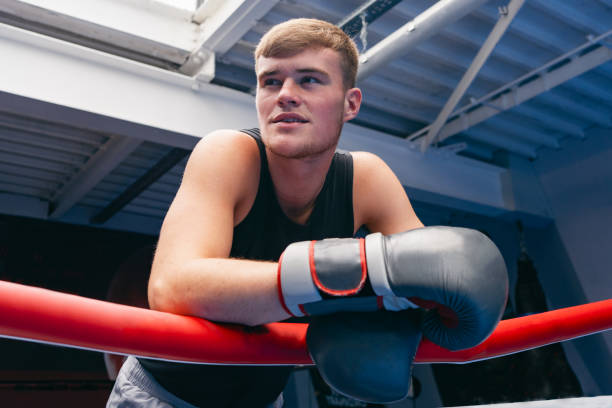 A young man wearing a sports vest and boxing gloves stands in a boxing ring. He leans on the ropes with a smile.
