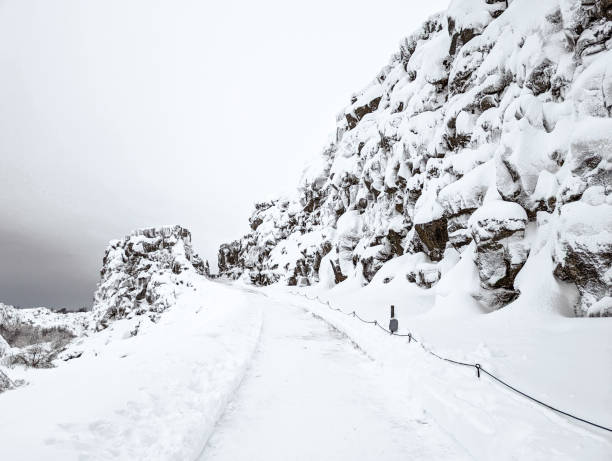 A snow-covered path winds through the dramatic rock formations in Thingvellir National Park, Iceland. The serene winter landscape highlights the rugged beauty and tranquil atmosphere of this historic national park.