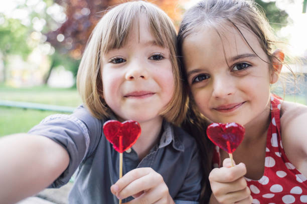 A boy in a grey shirt and a girl in a red polka-dot dress holding heart-shaped lollipops towards the camera, smiling warmly in a close-up selfie image taken in a sunny park.