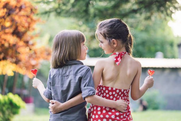 A young girl in a red polka-dot dress and a boy with long hair are seen from the back, holding red heart-shaped lollipops and embracing while sitting in a lush green park.