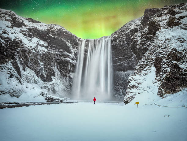 Northern Lights Over Skogafoss Waterfall in Iceland A person in a red jacket stands before the majestic Skogafoss waterfall, illuminated by the ethereal glow of the Northern Lights in Iceland. The snowy landscape and cascading water create a breathtaking scene of natural beauty and tranquility. skogafoss stock pictures, royalty-free photos & images