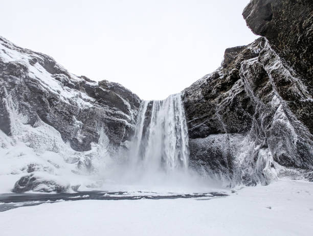 A stunning view of Skogafoss Waterfall in Iceland during winter. The waterfall flows powerfully between snow-covered cliffs, surrounded by an icy landscape that highlights the beauty of nature in the cold season.
