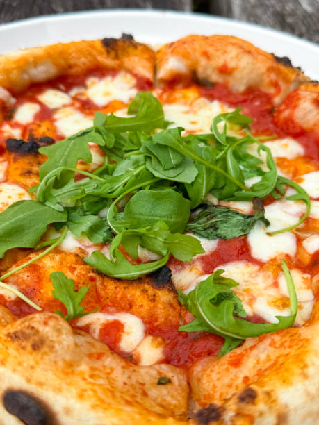 Full frame image of whole, freshly baked takeaway Margherita pizza on single-use disposable white plate, stringy, melted buffalo mozzarella cheese, rich tomato marinara sauce topping, garnished with rocket leaves, elevated view, focus on foreground