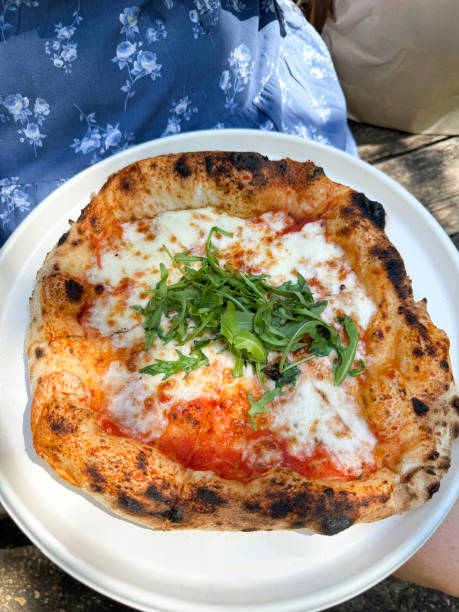 Image of whole, freshly baked takeaway Margherita pizza on single-use disposable white plate, stringy, melted buffalo mozzarella cheese, rich tomato marinara sauce topping, garnished with rocket leaves, wooden table background, elevated view
