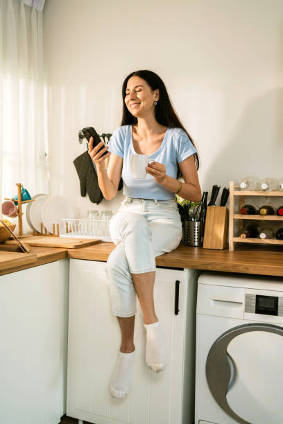 istock An attractive Caucasian woman using a smartphone while relaxing, drinking coffee and sitting on the kitchen counter at home in the morning. Happy woman shopping on mobile phone while in the kitchen. 2161296116