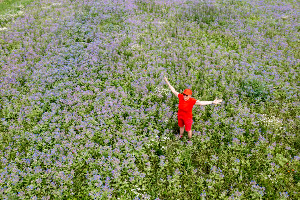A woman in red clothes and a cap relaxing, outstretching arms in a meadow of blue flowers seen from above in summer.