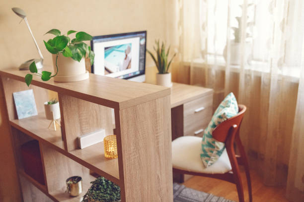 Cozy bright room with large window and workplace nearby. Light color wooden shelf with home plants, candles and boxes. Table with drawers and monitor on it and wooden chair with pillow on background.