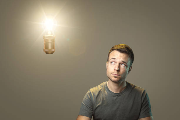 A man in a t-shirt is looking thoughtfully at a glowing light bulb suspended above him. Concept of having an idea or a moment of inspiration.