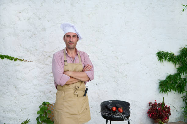 A confident chef wearing an apron and hat stands next to a grill with vegetables in an outdoor setting. He is crossing his arms and looking straight at the camera.