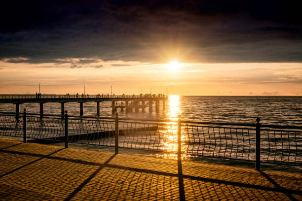 istock Orange sunset over the sea from under dark clouds. There is a long pier and a sunny path on the surface of the water. Long shadows from the fence of the embankment. 2161323783