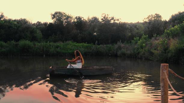 Woman in a white dress rows a boat on a lake and admires beautiful sunset, with the water reflecting the vibrant colors of the sky.