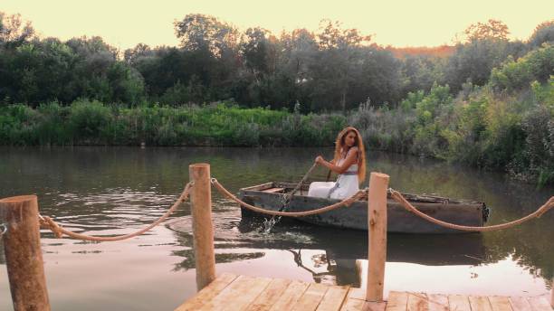 Woman in a white dress rows a boat on a serene lake at sunset, with the sky's reflection on the water.