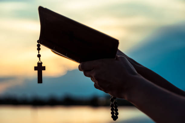 Hands holding an open Holy Bible with a wooden cross over sunset
