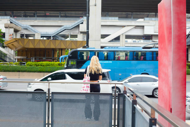 Young blond thai woman is standing at multilane road Vivhadi Rangsit Road on side with traffic ruling to south. Woman is wearing casual clothes and has a bag around shoulder with repeated text THAILAND. She is standing close to pedestrian footbridge to new Bang Khen station behind traffic and overpass highway. Scene is near exit of campus of Kasetart University.