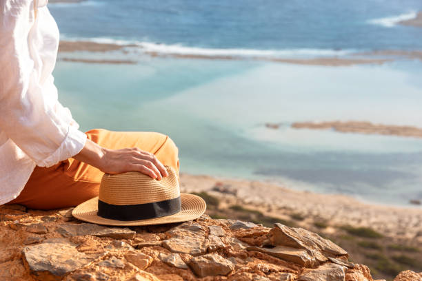 Person relaxing by the ocean, holding a straw hat on a rocky shore. Peaceful seaside moment with a scenic view of the water.