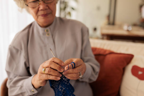 Charming elderly woman knitting while sitting on sofa at home.