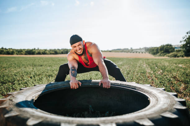 A young man is engaged in rigorous physical training by flipping a large truck tire at a park. This fitness activity takes place during a sunny morning, showcasing the individual's dedication and strength as he focuses on his workout routine amidst a vibrant outdoor setting.