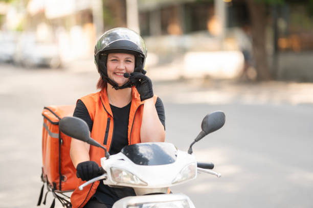 A female courier is seen riding a motorcycle on the road. She is smiling at the camera and holding a microphone in one hand. This scene reflects the dynamic and customer-focused aspect of modern delivery services.