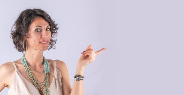 A middle-aged, short hair, brunette and beautiful woman look at the camera and pointing copy space on right side of the frame. Light purple background and large copy space.