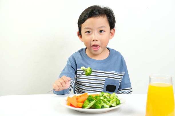 istock A young boy with a playful expression holds a fork with broccoli, sitting in front of a healthy meal and orange juice 2161415231