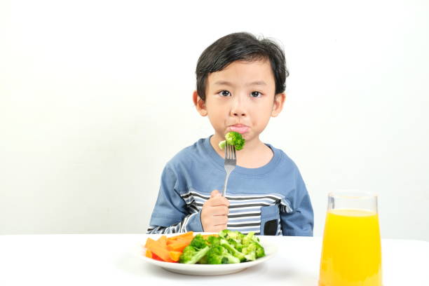 istock A young boy in a blue shirt pouts while eating broccoli from a fork, with a plate of mixed vegetables and a glass of orange juice. 2161415271