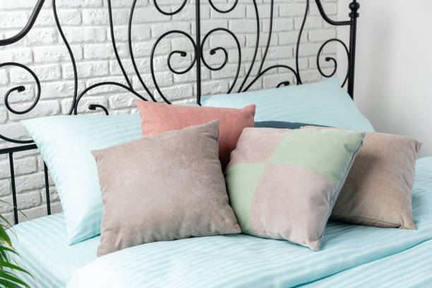 A close-up image of a bed with a blue sheet and colorful pillows, including pale pink, beige, and green.