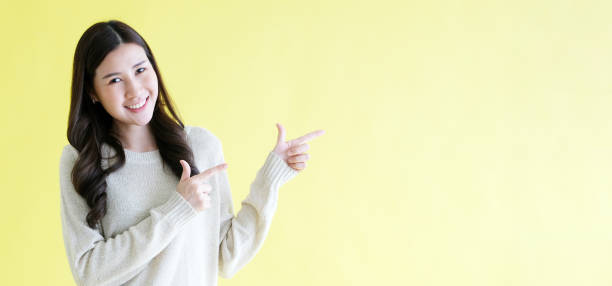 istock Happy young asian woman pointing hand for presenting product mock up, template while standing over isolated yellow background with copy space 2161434540