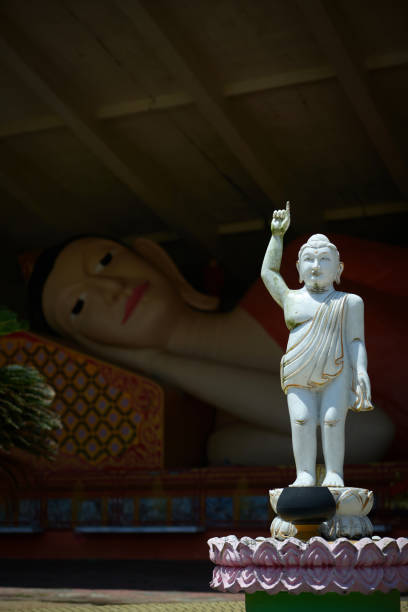 White young Buddha statue pointing a finger to the sky, with a sleeping Buddha statue in the background.