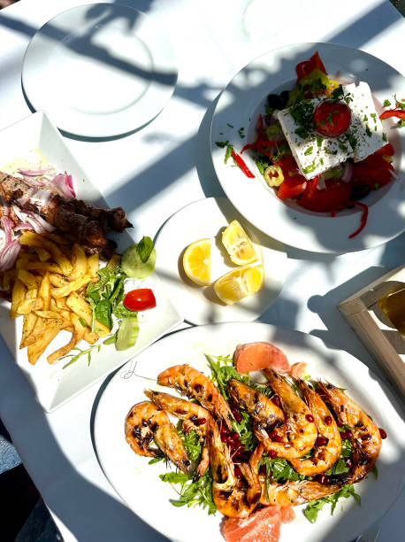 An appetizing set of traditional Greek dishes on the holiday table. In the foreground are luxurious grilled shrimp, a Greek salad with olives, feta, cucumbers, tomatoes and red onions, as well as juicy meat seasoned with aromatic herbs. The dishes are served on a wooden table decorated with greens and lemons, which creates the atmosphere of an authentic Greek feast. The photo conveys the spirit of Mediterranean cuisine and its rich flavors.