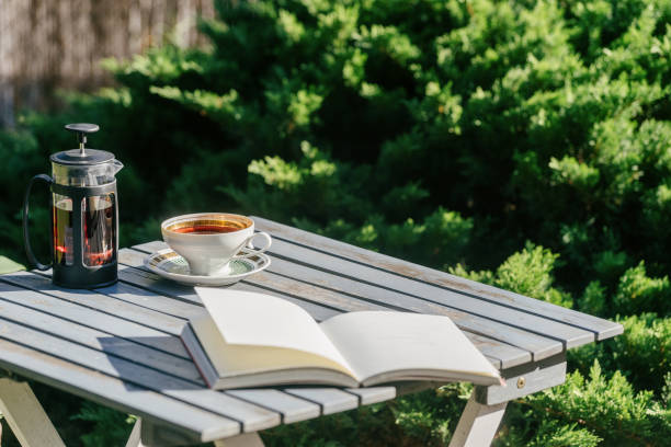 Wooden table with a cup of tea, teapot and an open book with copy space pages on garden table against green foliage. Summer backyard with place for reading and recreation on weekends