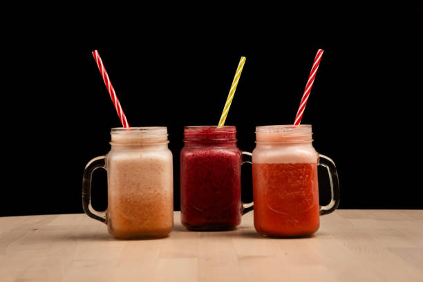 The three mason jar glasses filled with different smoothies and straws on a wooden table against black background