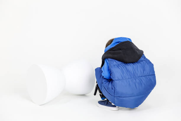 istock A child in a blue jacket and black pants crouches with head bowed, facing away from the camera. In front of him lies an upside-down white hourglass-shaped object. 2161453790