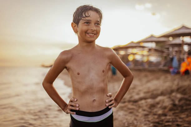A happy young boy with a playful smile standing by the beach during sunset, with hands on his hips, exuding a joyful and carefree vibe against a beautiful ocean backdrop.