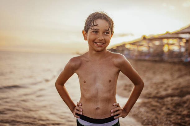 A cheerful young boy with a big smile enjoying his time by the beach during sunset, standing confidently with hands on his hips, highlighting joy and freedom in a serene setting.