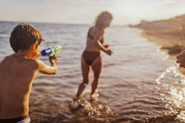 A young child aims a water gun at another kid while someone runs away, highlighting fun and excitement at the beach on a warm, sunny day.