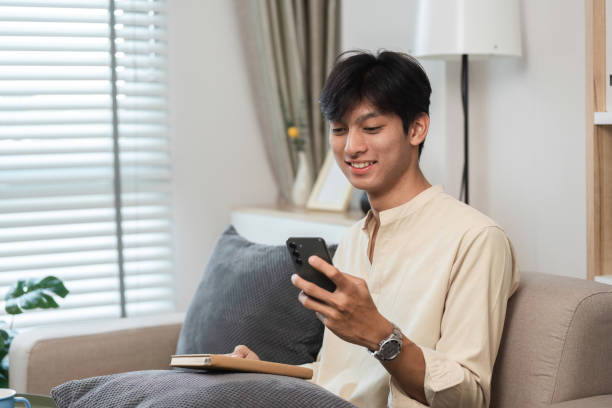 istock Young Man Relaxing on Couch in Modern Living Room, Using Smartphone and Holding Notebook, Casual and Comfortable Home Setting 2161454879