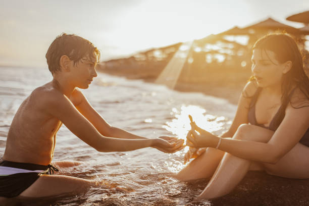 Two kids are sitting in the shallow water of a beach, playing with sand and each other, while the sun sets behind them, casting a beautiful glow over the scene.