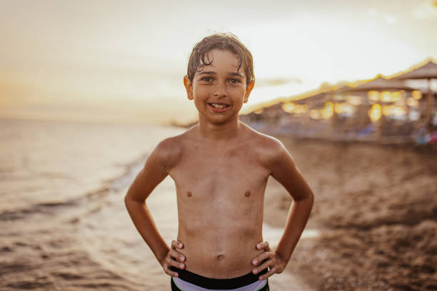 A smiling young boy enjoying his time at the beach during sunset, standing confidently with hands on his hips, perfectly capturing the essence of a joyful and carefree summer moment.